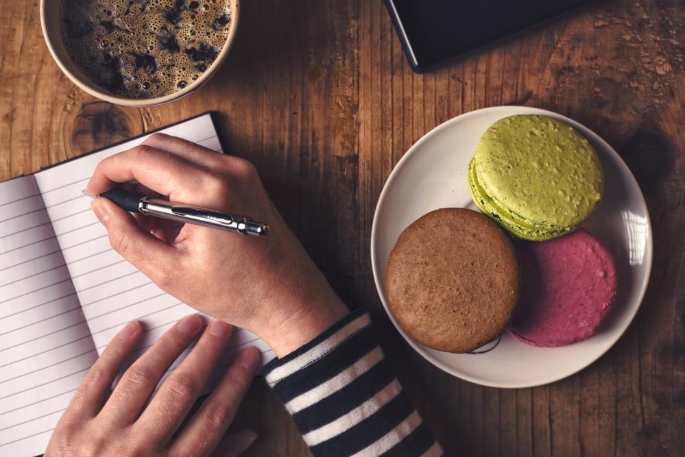 Image of young woman's hands writing in notebook with coffee and macaroons on the table.